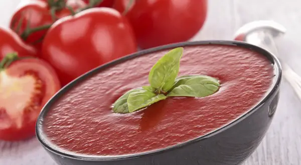 Un bol de soupe de tomates fraîches, garni de basilic, avec des tomates mûres en arrière-plan.