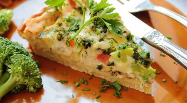 Une part de tarte aux légumes, garnie de persil frais, est servie sur une assiette orange avec du brocoli.