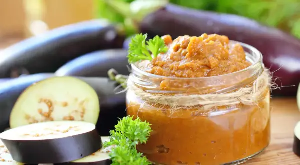 Un pot de caviar d'aubergine, prêt à être dégusté, avec des aubergines fraîches et des herbes. Une recette italienne simple et savoureuse.