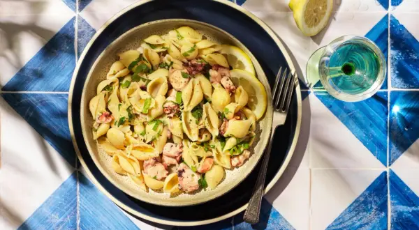 Un plat de pâtes aux coquillages et morceaux de poulpe, garni de persil frais et de quartiers de citron. Un verre de vin blanc et une fourchette complètent cette scène estivale.