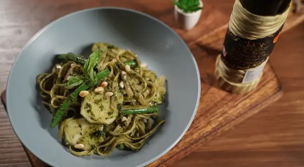Assiette de trenette al pesto, garnie de haricots verts, pignons de pin et basilic frais. Une bouteille d'huile d'olive est visible à côté.