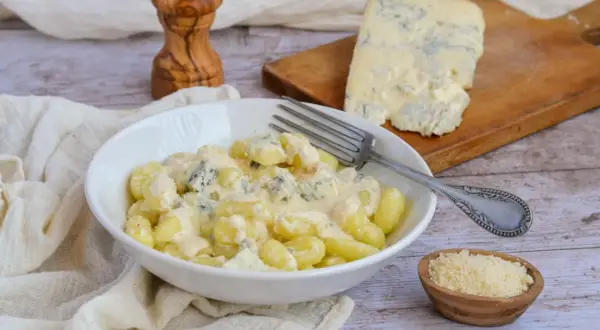 Gnocchi al gorgonzola crémeux dans un bol blanc, avec un morceau de gorgonzola sur une planche à découper en arrière-plan.