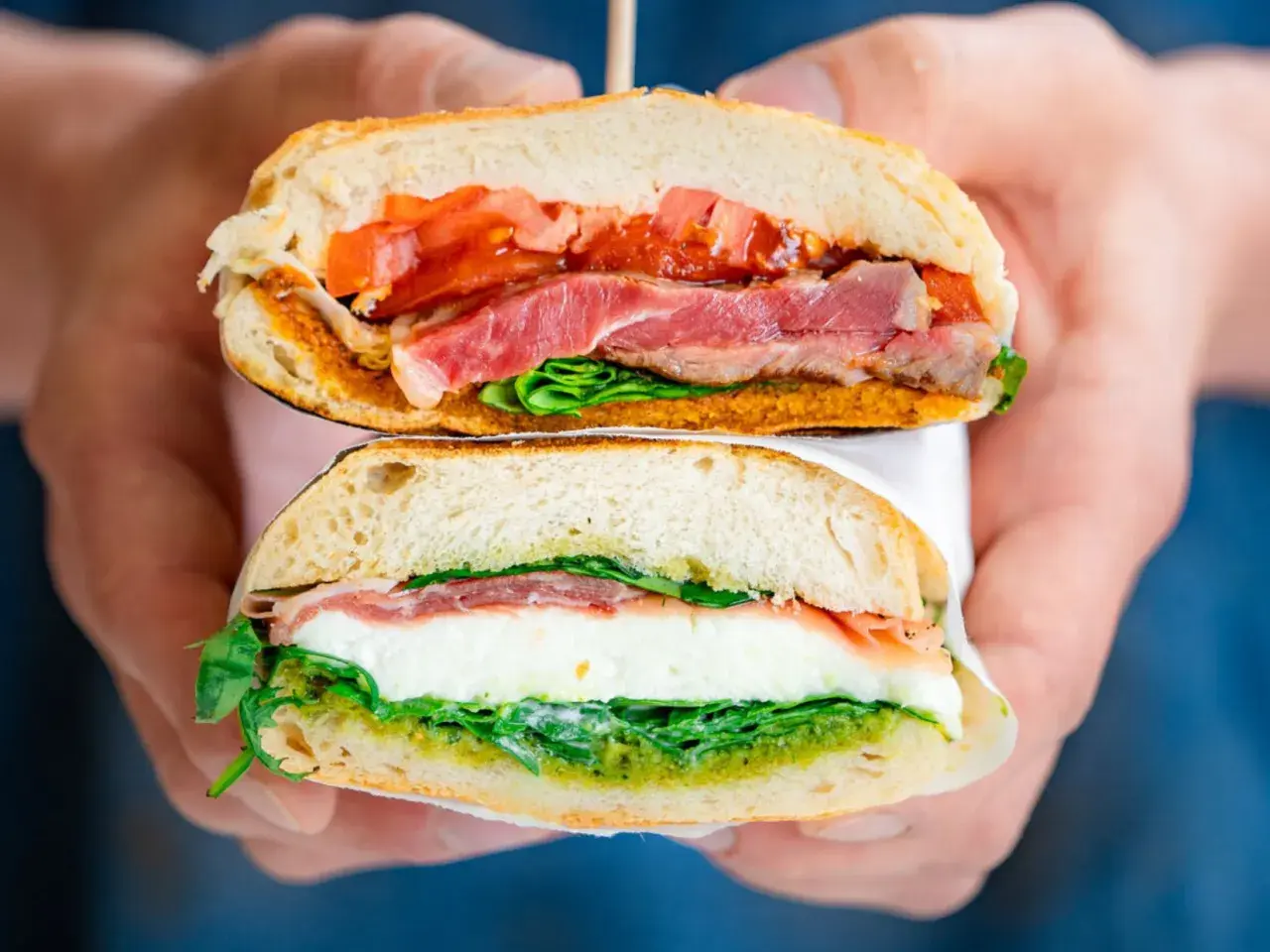 Deux moitiés d'un sandwich napolitain garni de jambon, mozzarella, tomates et pesto, tenues par des mains.