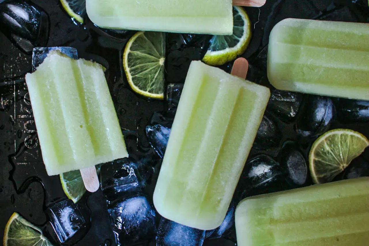 Plusieurs bâtonnets de glace au melon, avec des tranches de citron vert et des glaçons sur fond noir.