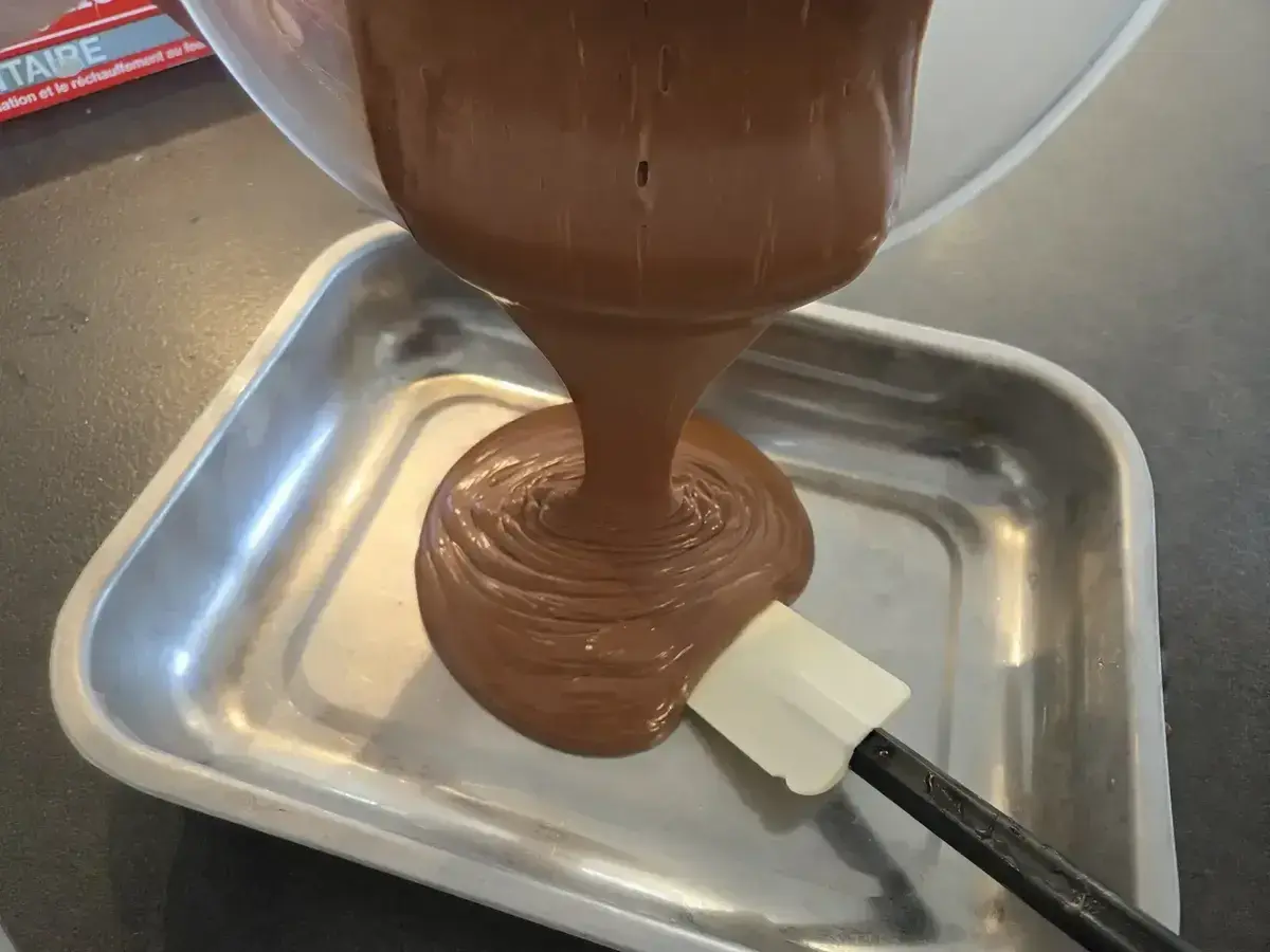 Une spatule aide à verser une ganache chocolat onctueuse dans un plat métallique.