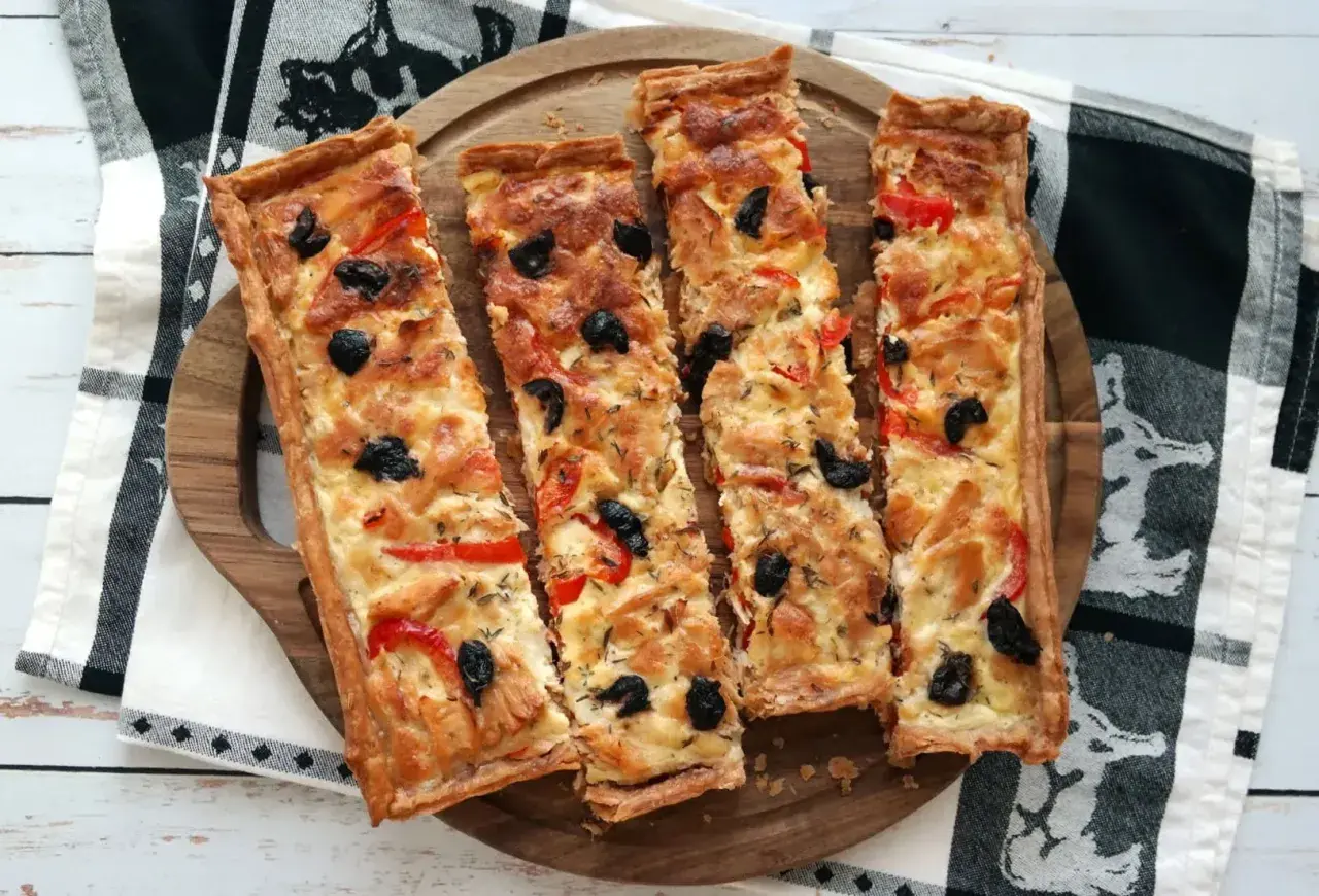Quatre parts de tarte au thon dorée, garnie d'olives noires et de poivrons rouges, sur un plateau en bois.