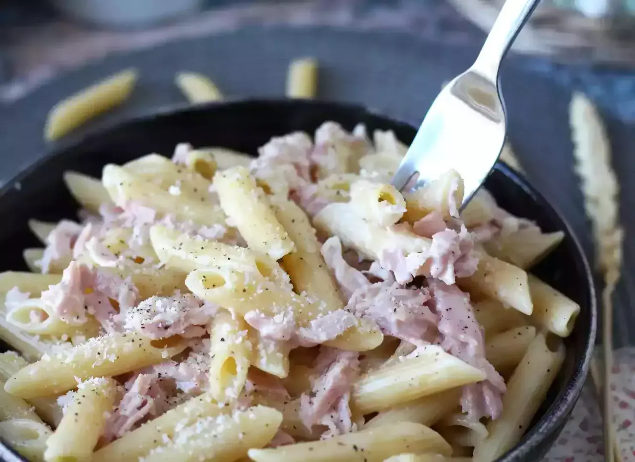 Une fourchette prélève des pâtes penne crémeuses avec du jambon effiloché, saupoudrées de parmesan et de poivre noir. Un plat de pâtes au jambon réconfortant.