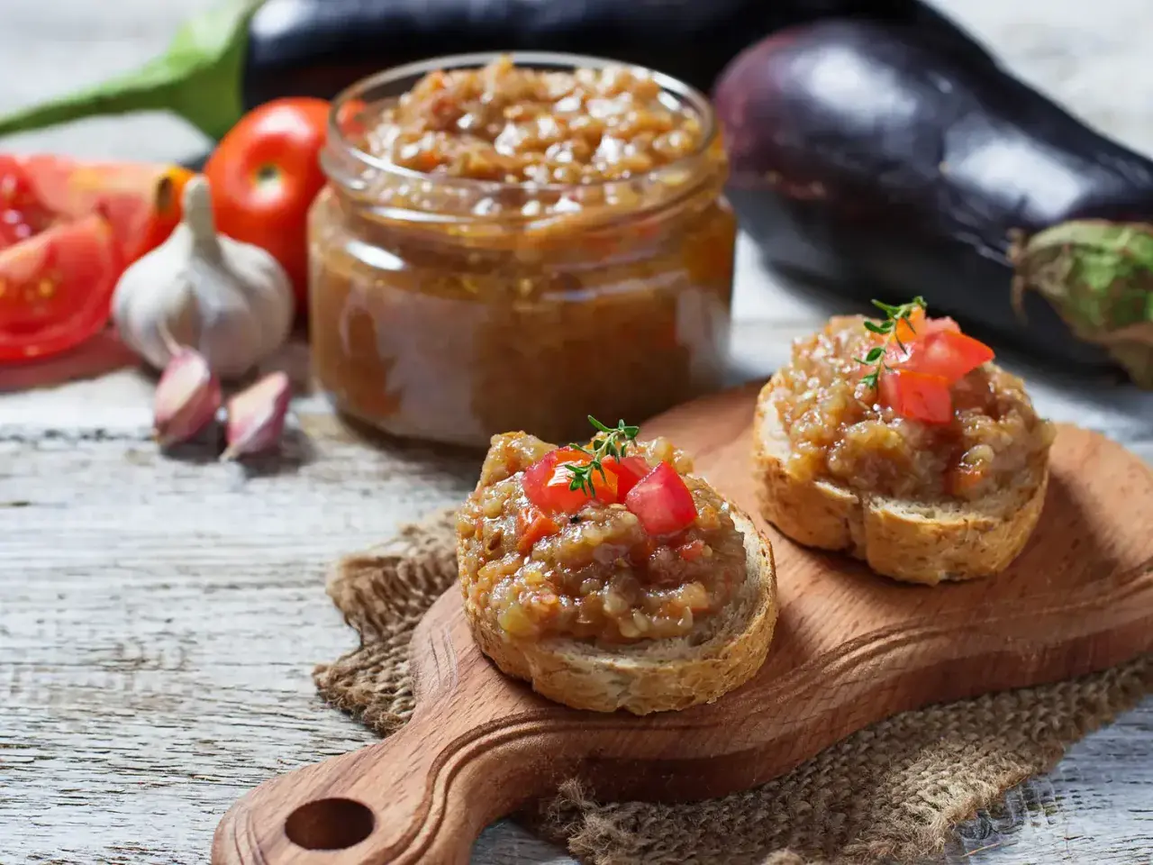 Deux toasts garnis de caviar d'aubergine, un pot de ce délice en arrière-plan, avec des aubergines, des tomates et de l'ail.
