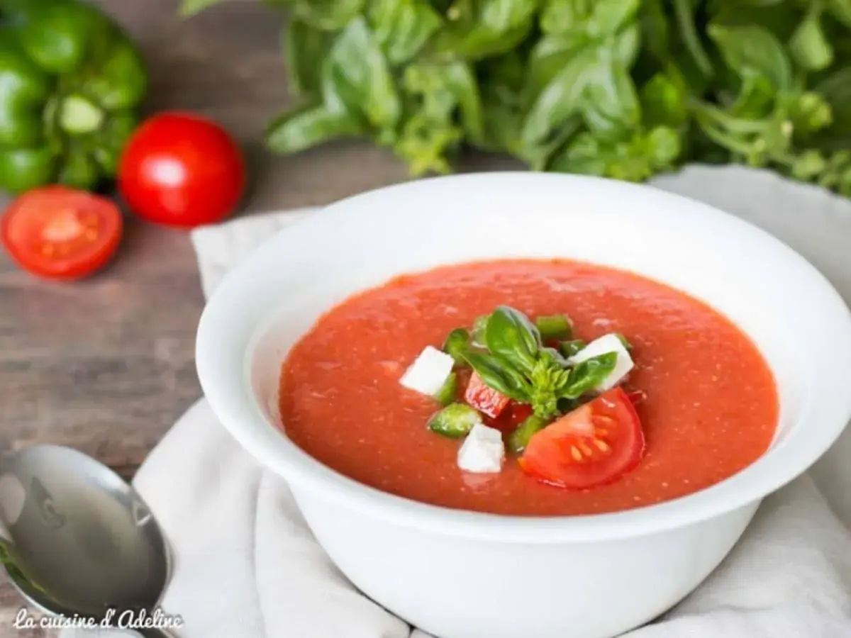 Un bol de gaspacho tomate frais garni de basilic, dés de poivrons et fromage. Des tomates, un poivron vert et du basilic frais en arrière-plan.
