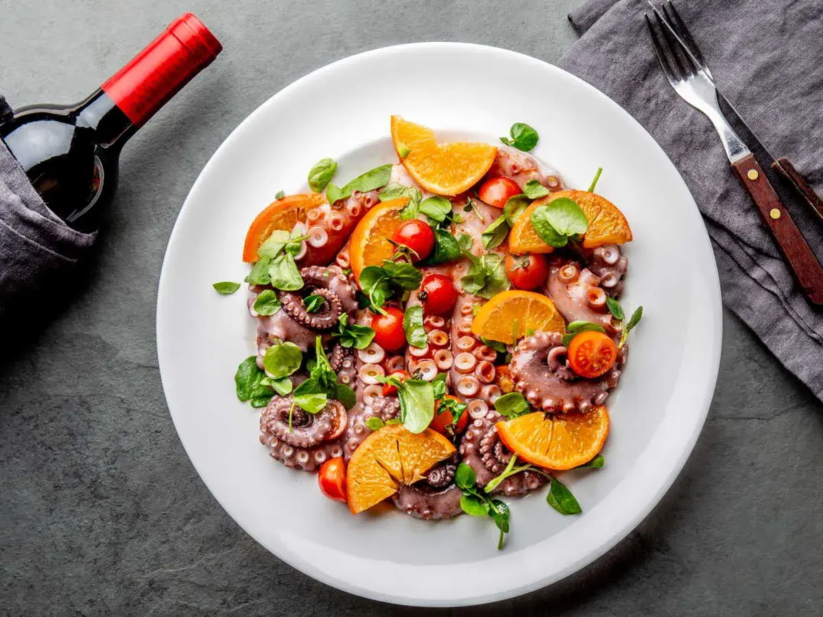 Salade de poulpe fraîchement préparée, garnie d'oranges, de tomates cerises et de verdure, prête à être dégustée avec une bouteille de vin rouge.