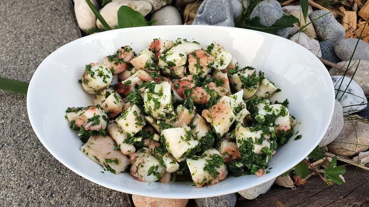 Salade de poulpe aux pommes de terre, assaisonnée d'herbes fraîches. Un délice méditerranéen servi dans un bol blanc, entouré de galets et de bois.