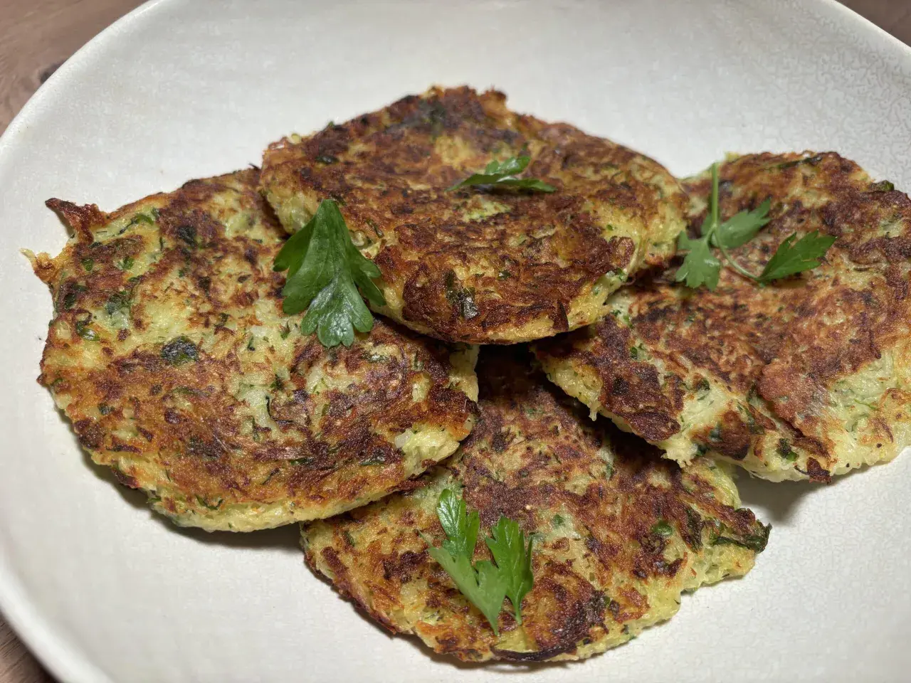 Galettes dorées de courgette et pomme de terre, garnies de persil frais, présentées dans un plat blanc.