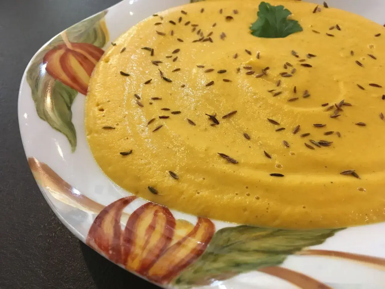 Velouté de carotte onctueux, parsemé de graines de cumin et garni d'une feuille de persil, servi dans une assiette décorée de motifs automnaux.