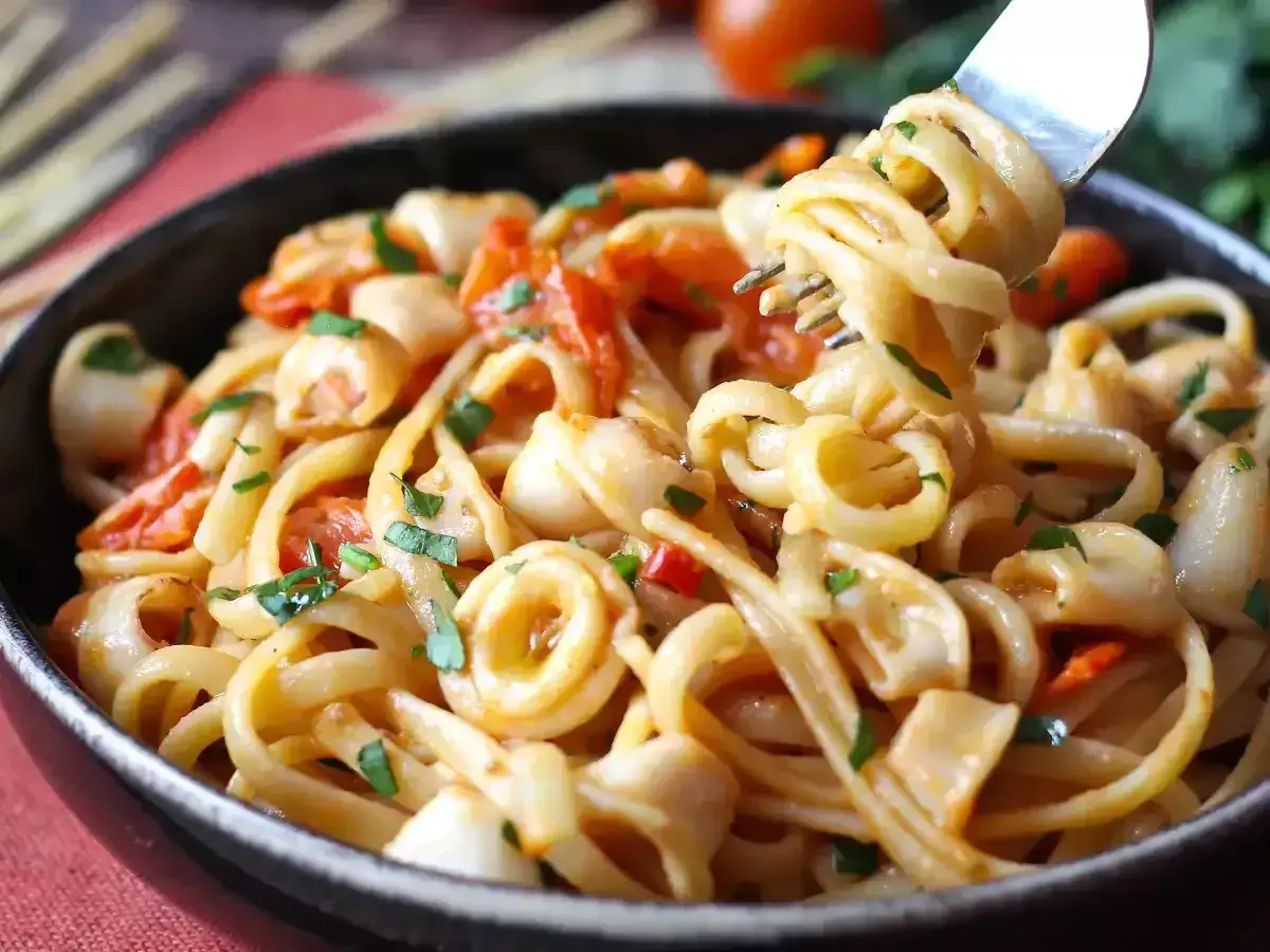Une assiette de pâtes aux calamars italienne, garnie de persil frais et de morceaux de tomates. Une fourchette soulève une portion de ce plat savoureux.
