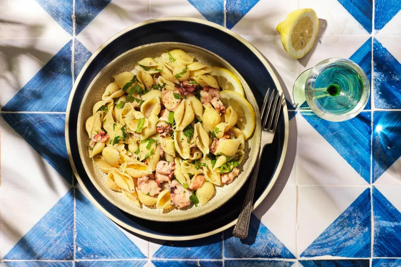Un plat de pâtes aux coquillages et morceaux de poulpe, garni de persil frais et de quartiers de citron. Un verre de vin blanc et une fourchette complètent cette scène estivale.
