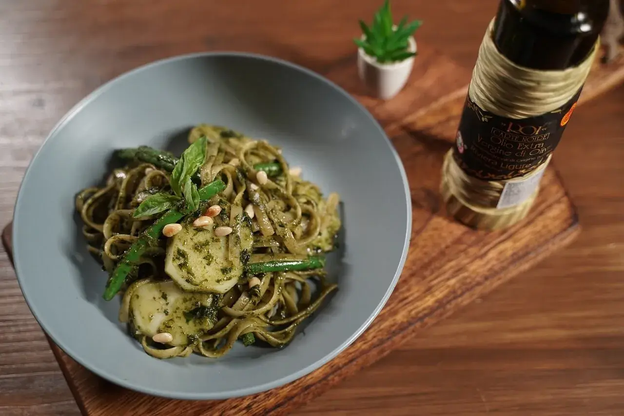 Assiette de trenette al pesto, garnie de haricots verts, pignons de pin et basilic frais. Une bouteille d'huile d'olive est visible à côté.