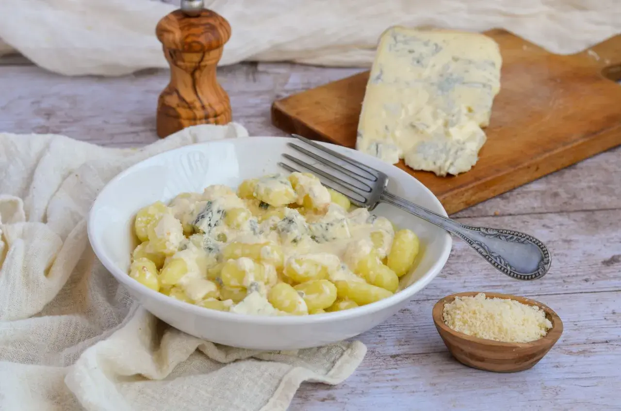 Gnocchi al gorgonzola crémeux dans un bol blanc, avec un morceau de gorgonzola sur une planche à découper en arrière-plan.