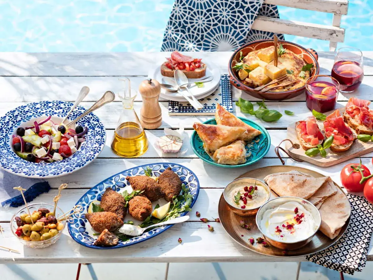 Idée apéro rapide sans cuisson : salade grecque, houmous, pain pita, feuilletés, bruschettas et olives, le tout près d'une piscine.