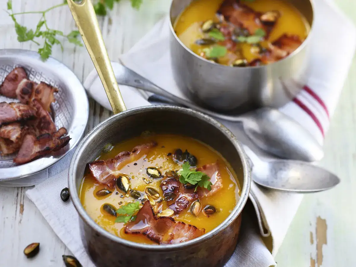 Soupe potimarron onctueuse garnie de lard croustillant et de graines de courge. Une assiette de lard fumé est à côté.