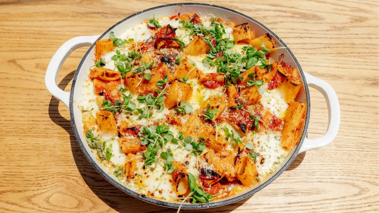 Plat de rigatoni alla sorrentina gratiné, garni de tomates, mozzarella fondue et herbes fraîches, dans une poêle blanche sur une table en bois.
