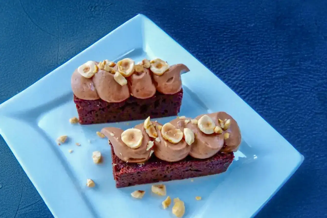 Deux d&eacute;licieux rectangles de brownie au chocolat, surmont&eacute;s d'une ganache chocolat a&eacute;rienne et de noisettes concass&eacute;es, sur une assiette bleue.