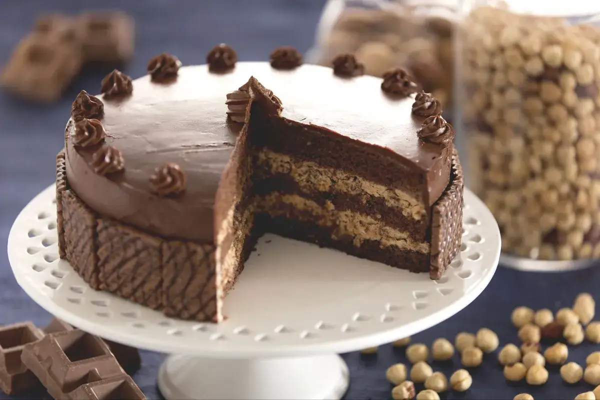 Gâteau gaufrette chocolat noisette, une tranche coupée révèle ses couches gourmandes. Des noisettes et des carrés de chocolat complètent cette œuvre.