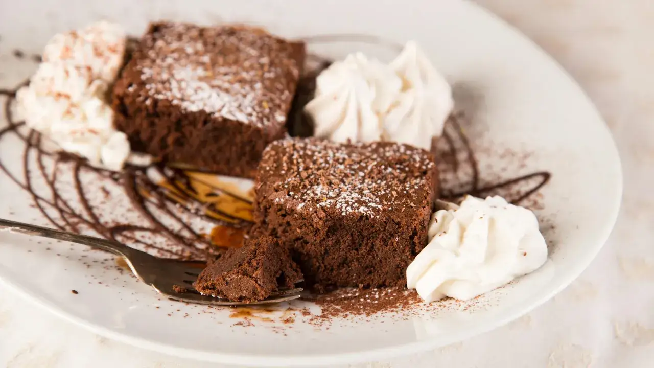 Deux parts de torta tenerina, saupoudrées de sucre glace et de copeaux de chocolat, accompagnées de crème fouettée sur une assiette blanche.