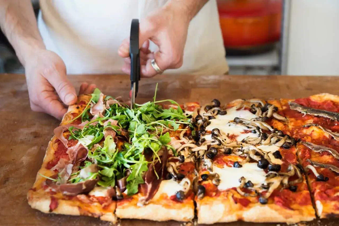 Une pizza romaine aux garnitures variées, dont du jambon cru et de la roquette, est coupée avec des ciseaux.