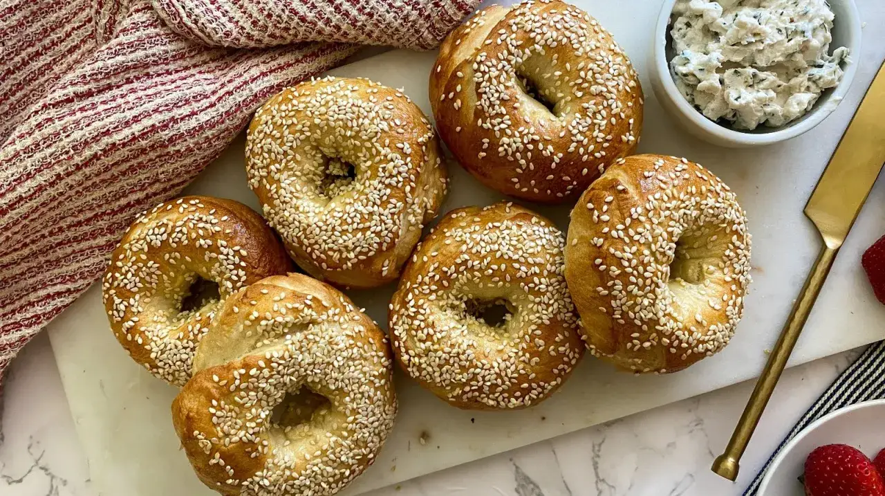 Six bagels garnis avec des graines de s&eacute;same, pr&ecirc;ts pour une recette bagel parfaite. Un bol de fromage &agrave; la cr&egrave;me et des fraises sont &agrave; proximit&eacute;.