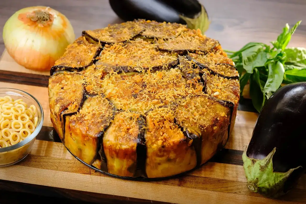Un plat rond et doré d'annelletti al forno, garni de tranches d'aubergine et de chapelure. À côté, des aubergines fraîches, un oignon et des pâtes annelletti.