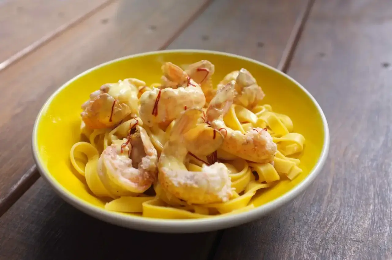 Un plat de p&acirc;tes aux langoustines cr&eacute;meuses, garni de fils rouges, servi dans un bol jaune vif sur une table en bois.
