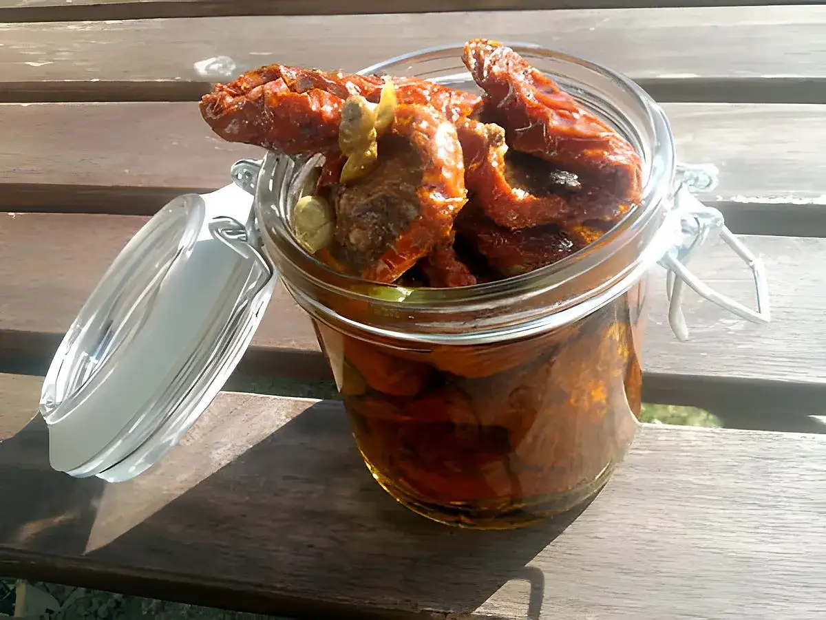 Un bocal en verre rempli de tomates séchées à l'huile, avec des herbes aromatiques. Le couvercle est ouvert sur le côté.