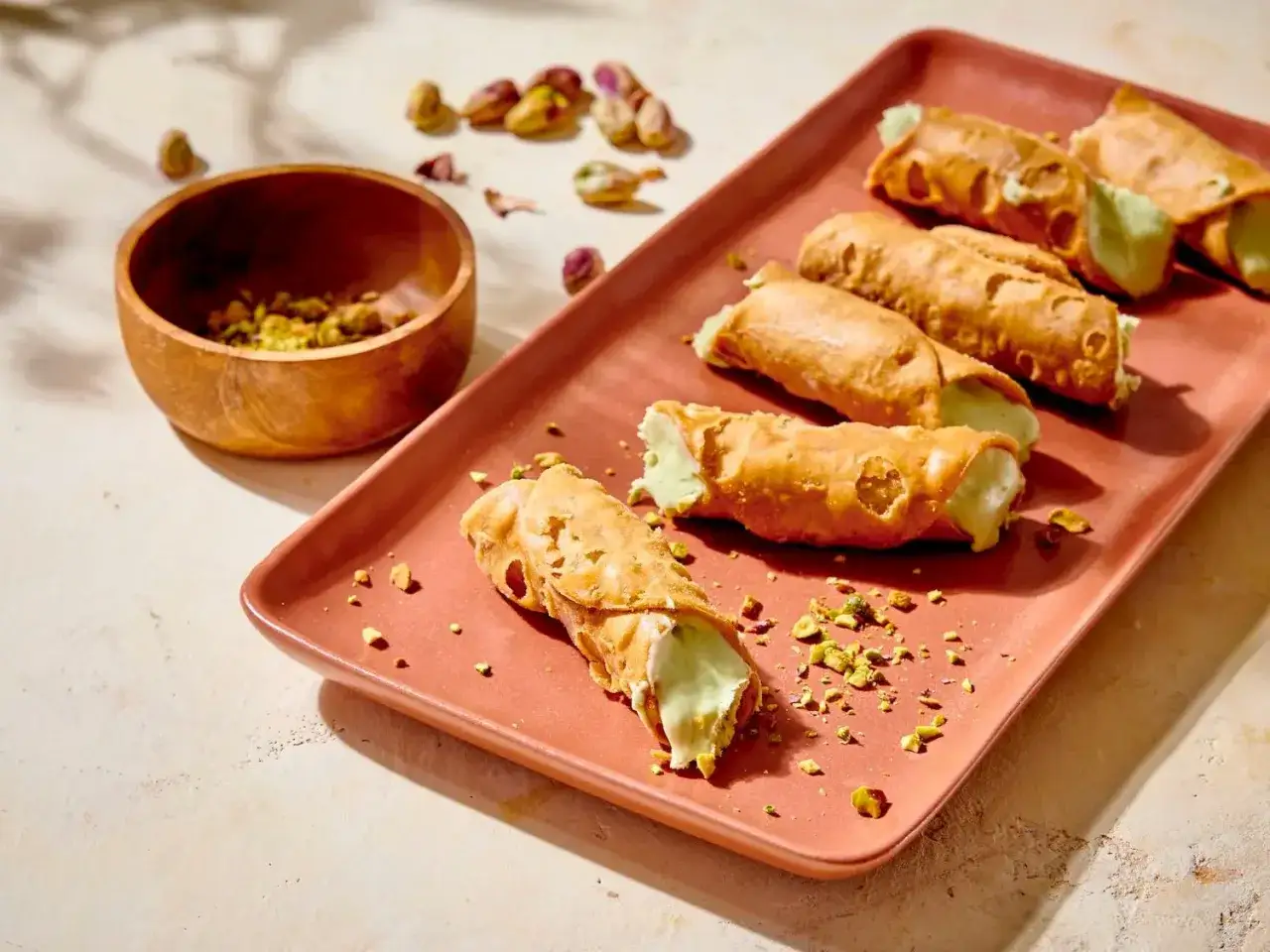 Cinq cannoli siciliens garnis d'une crème pistache, sur un plateau, avec des pistaches concassées et une coupelle en bois.