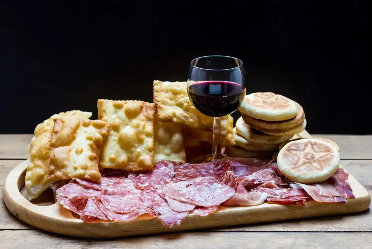 Plateau de charcuterie italienne avec du gnocco fritto croustillant, des pains ronds et un verre de vin rouge.