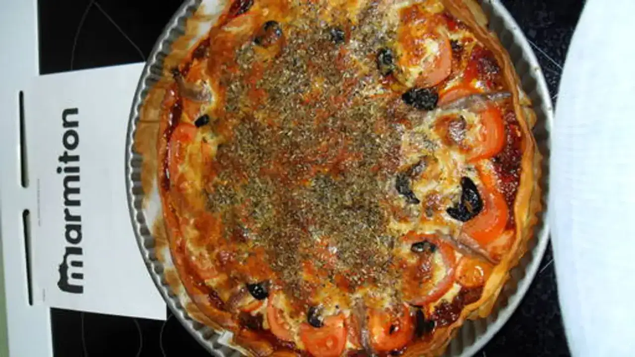 Tarte au thon garnie de tomates, d'olives et d'anchois, saupoudrée d'herbes.
