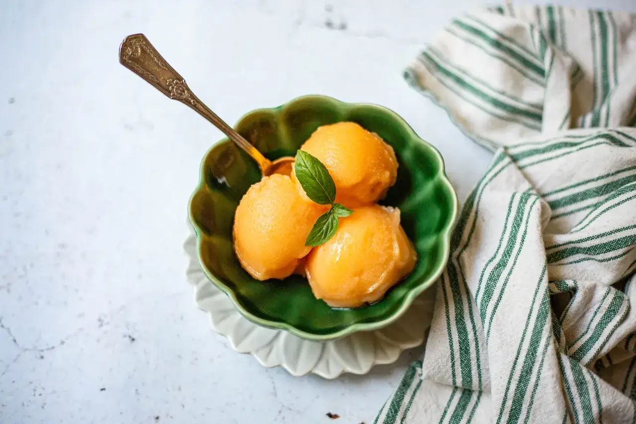 Trois boules d'une délicieuse glace au melon, garnies d'une feuille de basilic, dans un bol vert. Une cuillère antique est posée dessus.