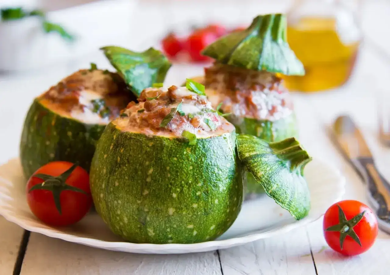 Trois courgettes rondes farcies, garnies de persil frais, sur une assiette blanche avec des tomates cerises et une bouteille d'huile d'olive en arrière-plan.
