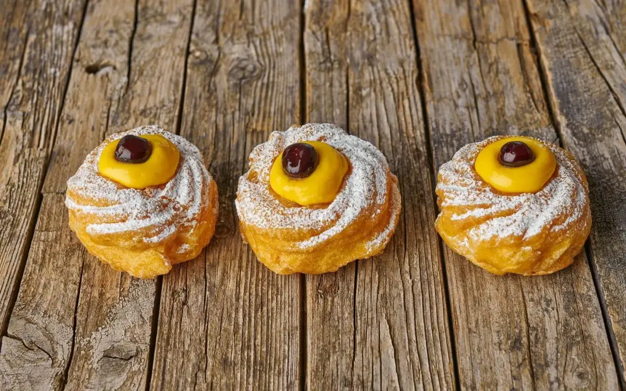 Trois zeppole di san giuseppe, pâtisseries italiennes saupoudrées de sucre glace, garnies de crème jaune et d'une cerise, sur fond de bois.