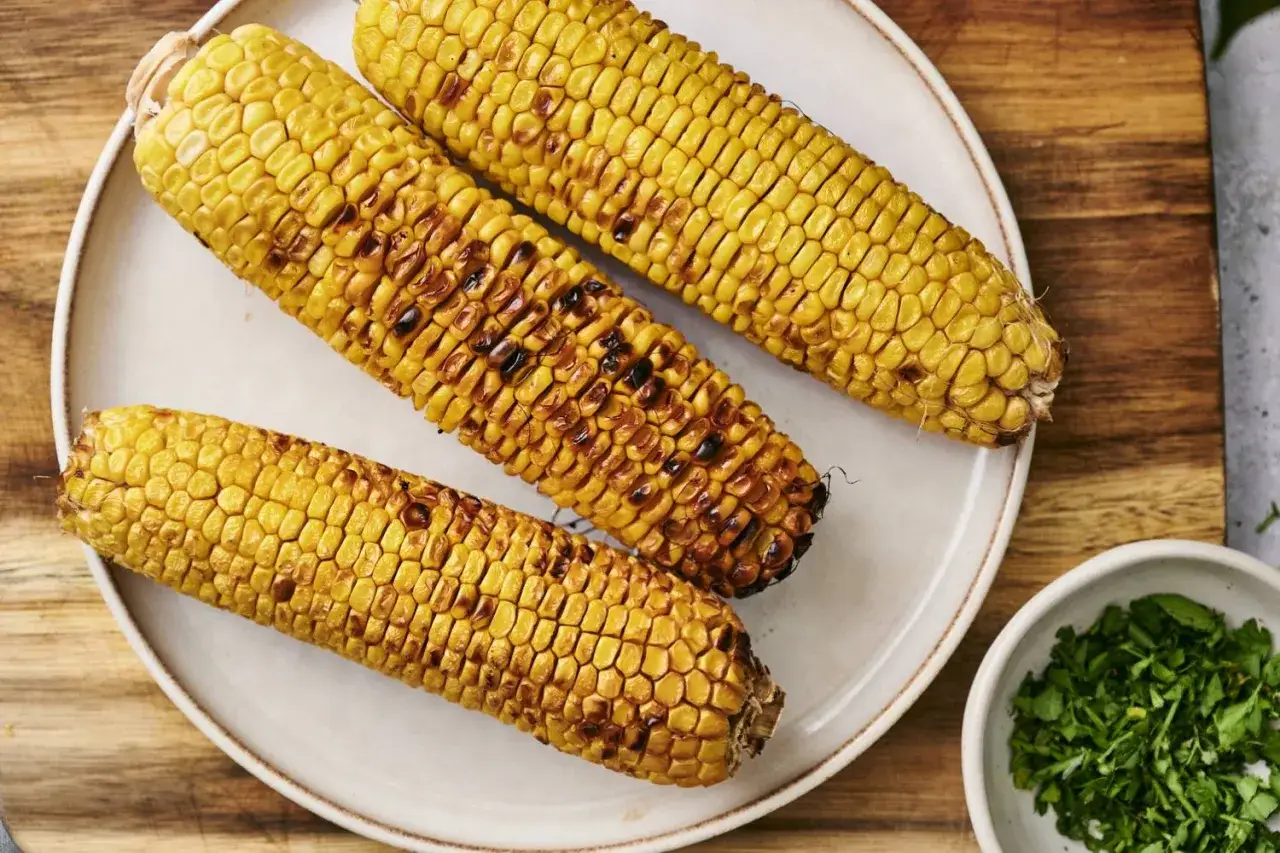 Trois épis de maïs grillé, dorés et légèrement caramélisés, sur une assiette blanche. Un petit bol de persil haché est à côté.