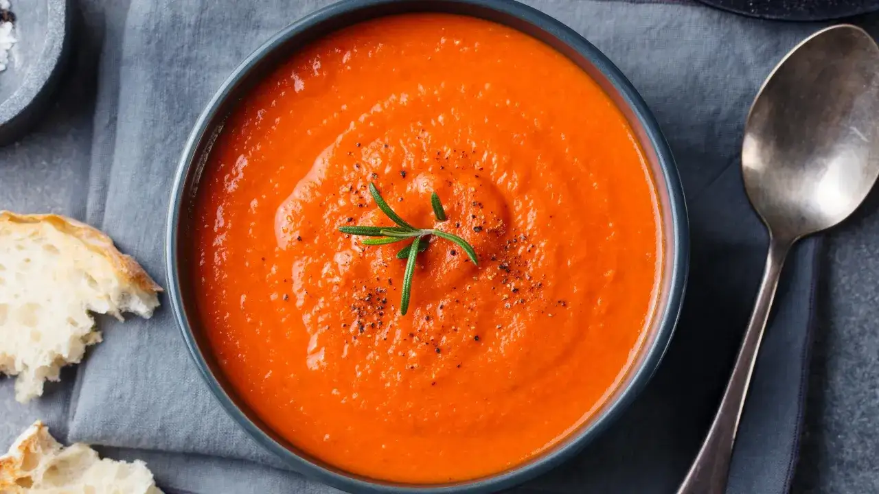 Un bol de soupe tomates fraîches, garni d'une branche de romarin et de poivre noir, servi avec du pain croustillant.