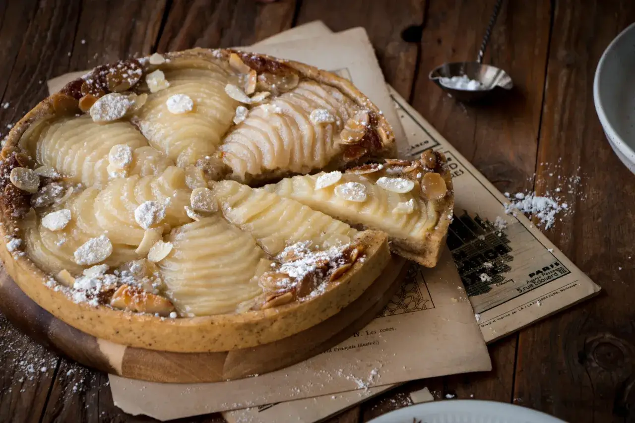 Une délicieuse tarte amandine aux poires, saupoudrée de sucre glace et d'amandes effilées, repose sur de vieux papiers.