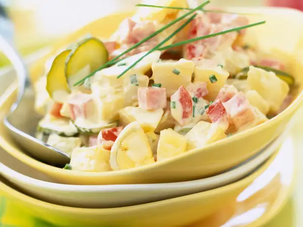 Salade piémontaise crémeuse avec pommes de terre, jambon, œufs durs, cornichons et ciboulette, servie dans un bol jaune.