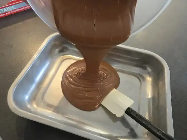 Une spatule aide à verser une ganache chocolat onctueuse dans un plat métallique.