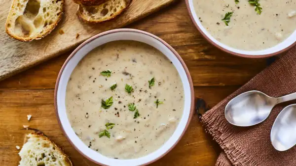Deux bols de velouté de champignons à la crème, garni de persil et de poivre, avec des tranches de pain croustillant sur une planche en bois.