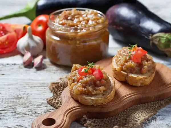 Deux toasts garnis de caviar d'aubergine, un pot de ce délice en arrière-plan, avec des aubergines, des tomates et de l'ail.