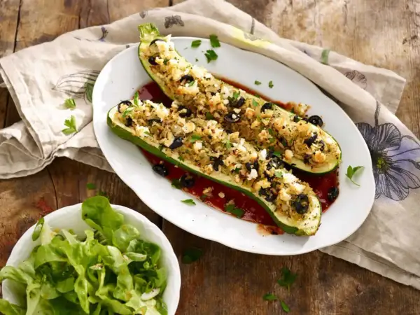 Deux courgettes farcies végétariennes, garnies de riz, olives et feta, sur un lit de sauce tomate. Une salade verte accompagne ce plat.