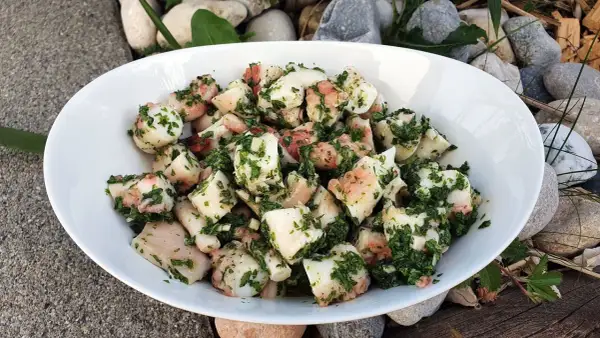Salade de poulpe aux pommes de terre, assaisonnée d'herbes fraîches. Un délice méditerranéen servi dans un bol blanc, entouré de galets et de bois.