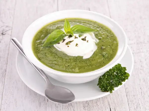 Soupe froide courgette fromage frais, garnie de basilic et persil. Une cuillère repose à côté du bol.