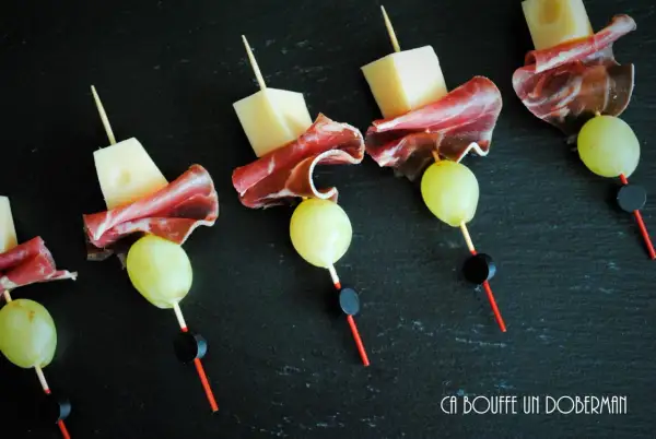 Délicieuses brochettes apéro : cubes de fromage, fines tranches de charcuterie et raisins verts sur fond noir.