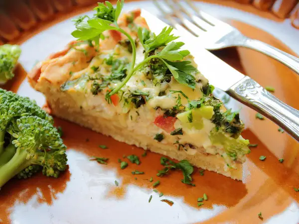 Une part de tarte aux légumes, garnie de persil frais, est servie sur une assiette orange avec du brocoli.